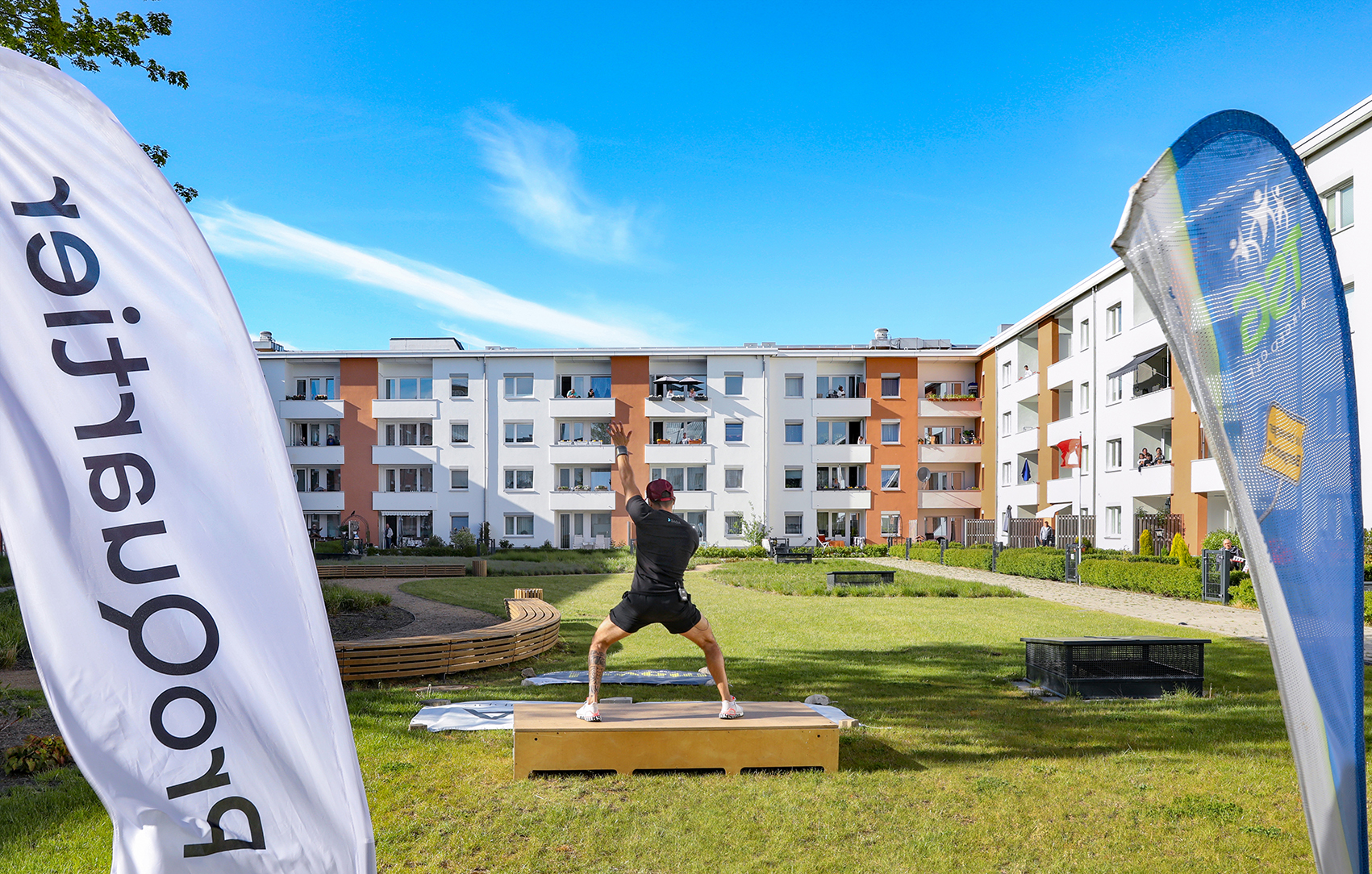 Ein Sporttrainer steht in Squathaltung auf einem Holzpodest und zeigt Sportübungen, im Hintergrund sind Wohngebäude