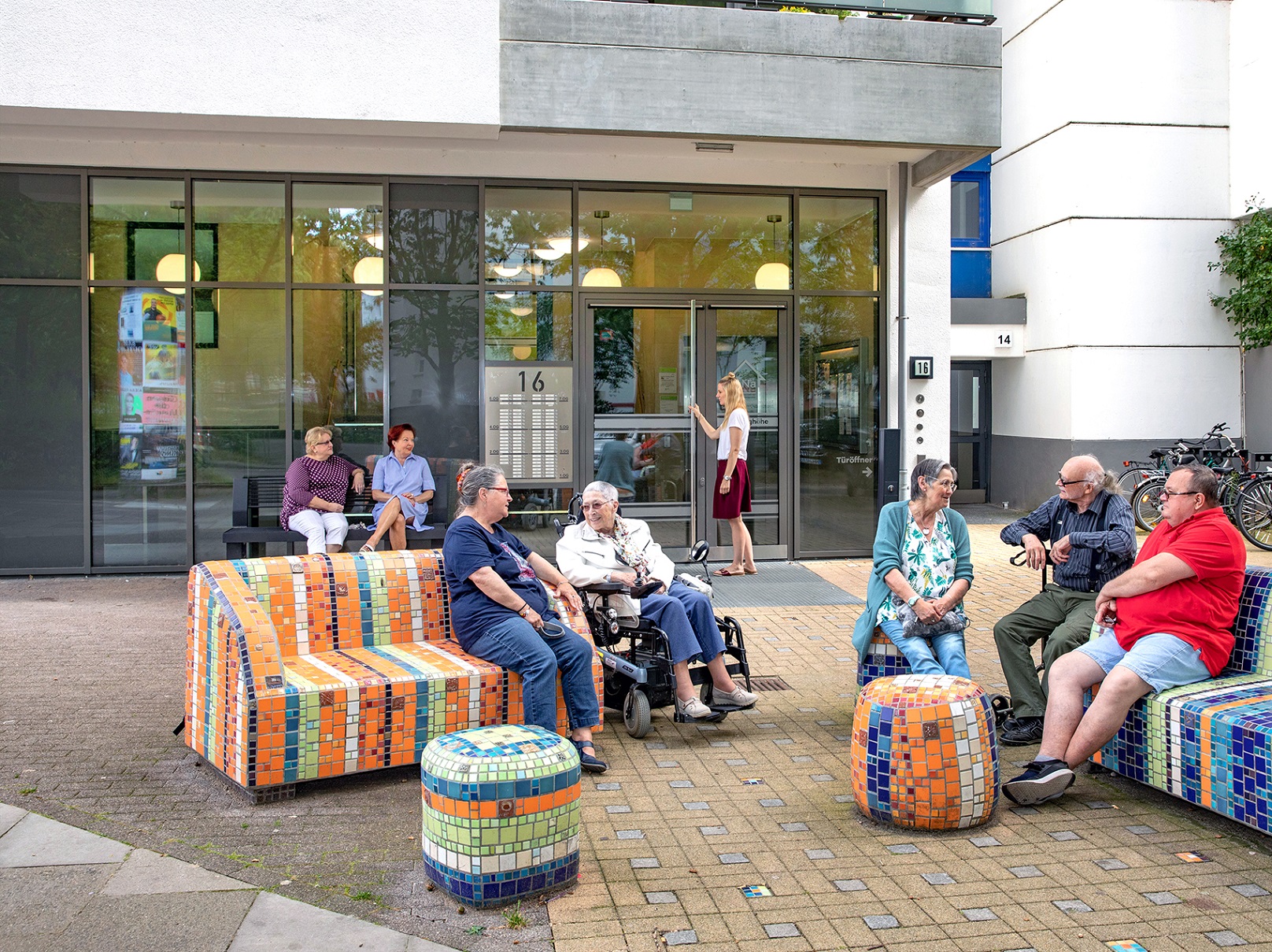 Eine Gruppe von Menschen sitzt vor einem Wohngebäude auf Sauf künstlerisch gestalteten öffentlichen Sitzgelegenheiten.