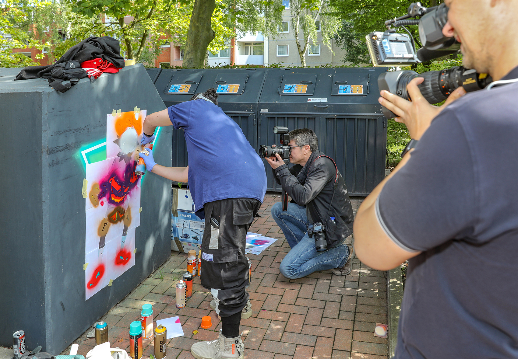 Ein Künstler sprüht ein Kunstwerk auf einen Müllstand  zwei ein Kamerateam fotografiert und filmt diesen dabei. 
