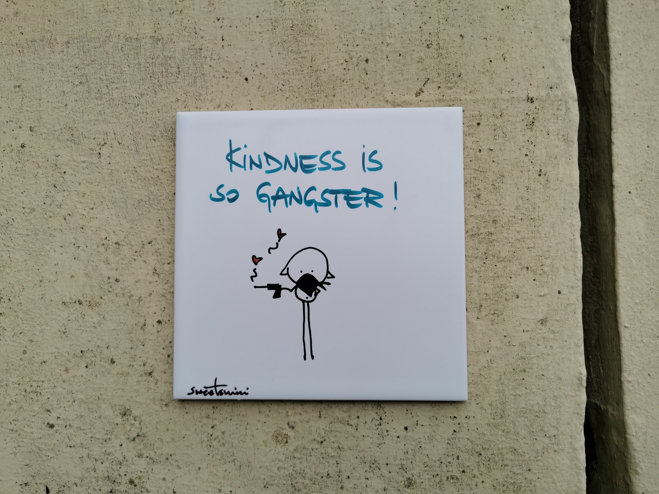 Eine künstlerisch gestaltete quadratische Kachel angebracht an einer Hausmauer. Auf der Kachel steht der Slogan "Kindness is so Gangster!" Darunter ist ein Strichmännchen, welches eine Pistole hält aus der Herzen geschossen kommen.