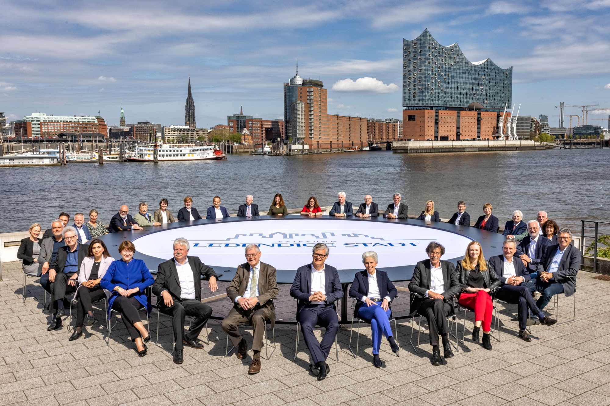 Foto © von Thorge Huter, darauf zu sehen ein großer runder Tisch unter freiem Himmel an dem 34 Personen sitzen , im Hintergrtergrund ist die Hafencity zu sehen