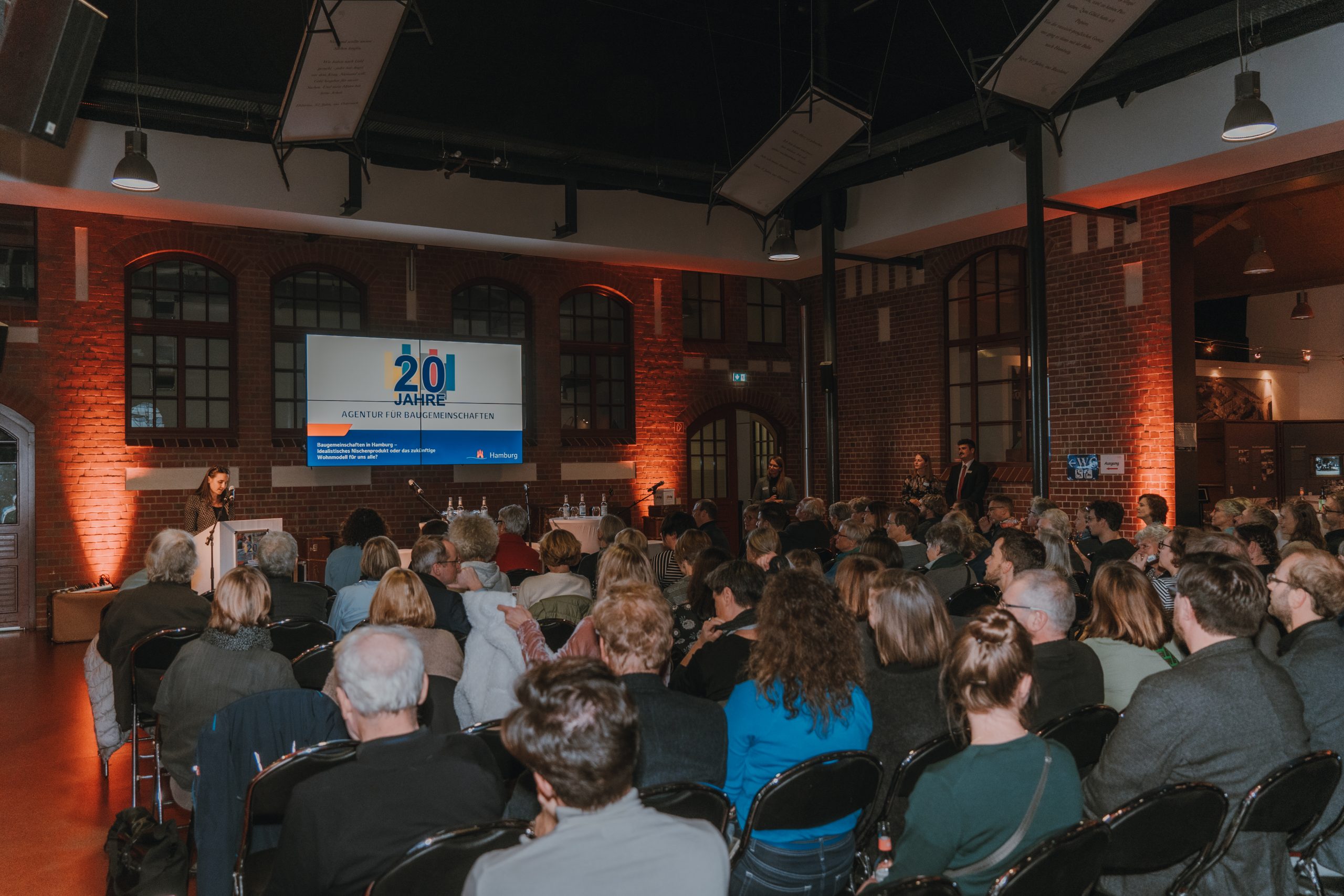 Saal mit Publikum und Rednerin vorne neben einem großen Bildschirm mit dem Titel "20 Jahre Agentur für Baugemeinschaften"