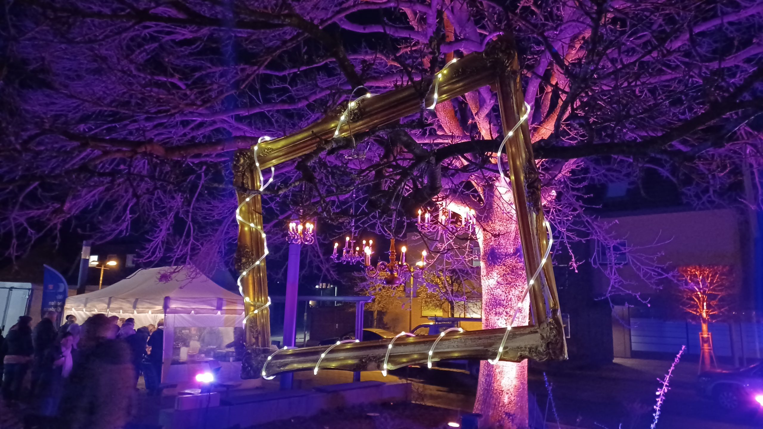 Farbig angeleuchteter Baum mit aufgehängten Kronenleuchtern und einem großen Bilderrahmen.