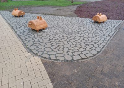 Gepflasterte Fläche an einem Fußweg mit hölzernen Spielelementen in Gestalt von Schweinen