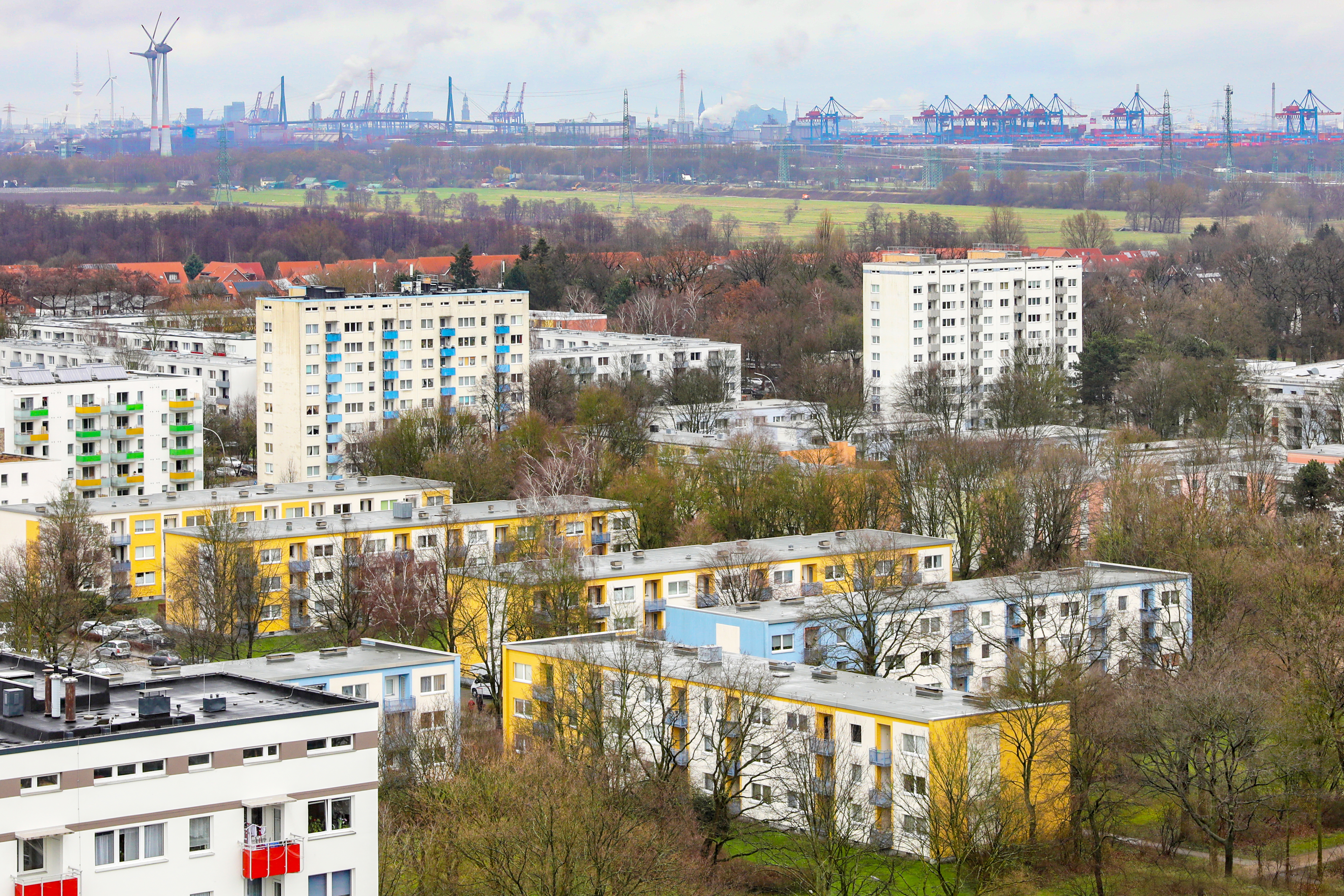 Luftaufnahme von einem Wohnquartier mit Mehrfamilienhäusern mit gelber und blauer Fassade, im Hintergrund Kräne vom Hamburger Hafen. 