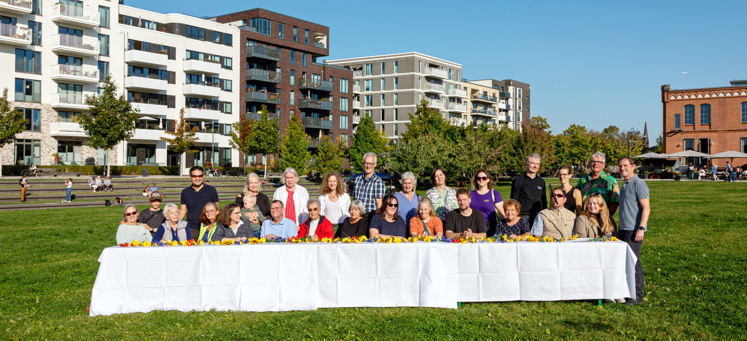 Gruppenfoto im Park an einer langen Tafel mit Gebäuden im Hintergrund.