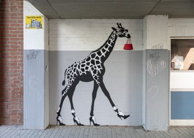 Streetart Kunstwerk an einer Wand. Zusehen ist eine schwarz weiße Giraffe, welches auf Stöckelschuhen läuft und im Maul eine rote Handtasche trägt.