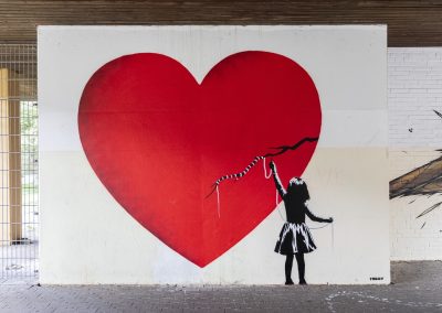 Streetart Werk an einer Wand. Zu sehen ist ein großes rotes Herz, mit einem Riss, welches ein kleines Mädchen wieder zusammen näht.